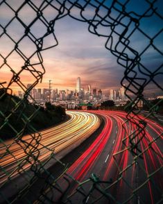 a city skyline seen through a chain link fence at dusk with the lights of cars streaking by