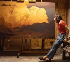 a man sitting in front of a large painting with clouds on the wall behind him