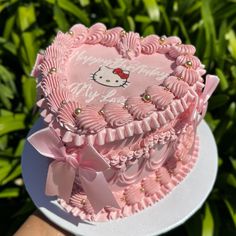 a hello kitty cake with pink icing and bows on it's top, sitting in front of some bushes