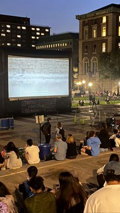 people sitting on the ground watching a movie in front of a large screen at night