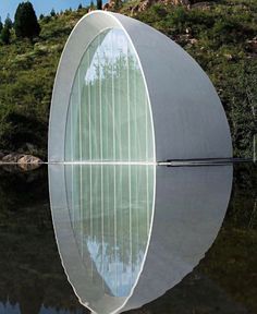 a circular structure sitting on top of a lake next to a lush green hillside covered in trees