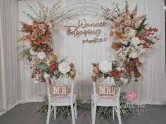 two white chairs with flowers on them in front of a backdrop