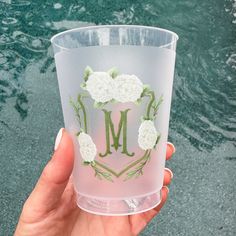 a hand holding up a clear cup with flowers on the bottom and letter m painted on it