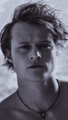 a shirtless young man wearing a necklace and looking at the camera while standing in front of water