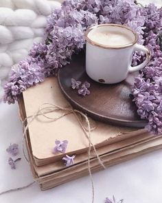 a cup of coffee sitting on top of a wooden plate next to some purple flowers