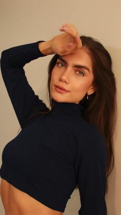 a woman with long hair wearing a black top and posing in front of a white wall