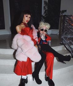 two women in red dresses and fur coats sitting on steps with one woman holding a cell phone