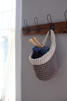 a crocheted basket hanging on the wall with two knitting needles and yarn in it