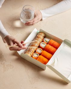 two people holding chopsticks near a box of sushi and rolls with water