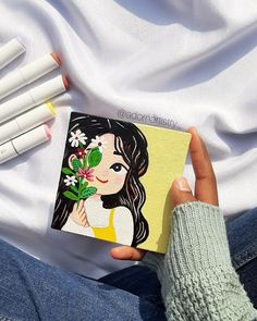 a person holding a small card with flowers on it next to some pens and markers