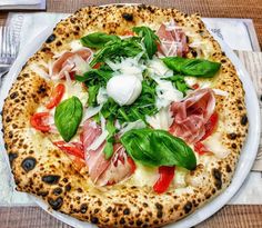 a pizza on a white plate topped with meat and veggies next to utensils