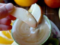 a person dipping an apple into a bowl of dip with oranges in the background