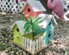 three colorful bird houses sitting on top of a gravel covered ground next to a pink flamingo