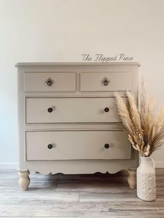 a white dresser sitting on top of a wooden floor next to a vase filled with dry grass