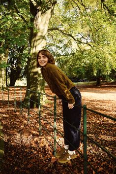 a woman leaning on a fence in the park with leaves all around her and smiling at the camera