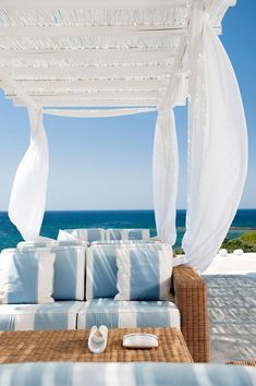 a bed sitting on top of a wooden table next to the ocean with white drapes