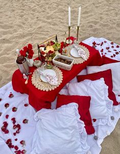 a table set up on the beach with red and white linens, flowers, candles and food