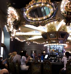 a group of people sitting at a bar with lights on the ceiling and mirrors above them