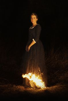 a woman in a black dress standing next to a fire with her hands on her hips