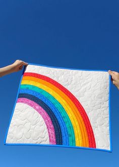 two hands holding up a quilted rainbow against a blue sky