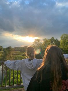 two people standing on a bridge with the sun setting in the background and clouds above them