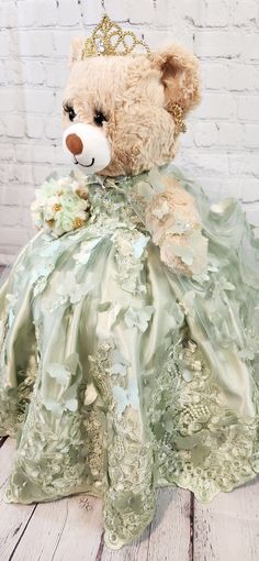 a teddy bear dressed in a green dress with flowers on it's chest and wearing a tiara