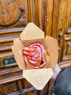 a person holding up a hot dog in a cardboard box with bacon on it and ketchup