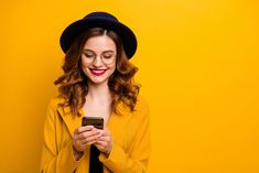 a woman wearing glasses and a hat looking at her cell phone while standing against a yellow background