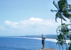 a man standing on top of a rock next to the ocean with palm trees in front of him