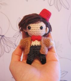 a hand holding a tiny crocheted doll in it's right hand and wearing a red hat