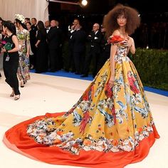 a woman in an orange and yellow dress standing on a white carpet with other people