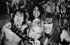 four shirtless men posing with guitars in front of graffiti covered walls and one holding his mouth open