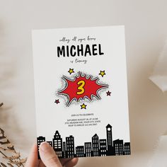 a hand holding up a birthday card with the number 3 on it, in front of a cityscape