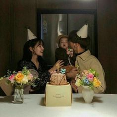 a group of people standing around a table with a cake in front of them and two vases filled with flowers