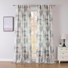 the curtains are hanging in front of a window with an artistic pattern on it, along with a white dresser and lamp