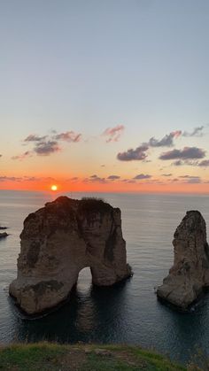 the sun is setting over two large rocks in the ocean near some cliffs and water