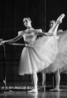 two ballerinas in white tutus and ballet shoes, one holding the bars