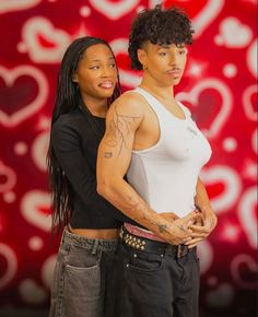 two people standing next to each other in front of a red wall with hearts on it