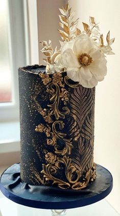 a black and gold wedding cake with white flowers on top is sitting on a blue stand