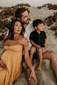 a man, woman and child are sitting on the sand in front of some plants