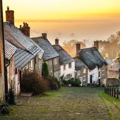 a cobblestone street with houses on both sides and fog in the background at sunset