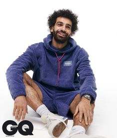 a man sitting on the ground wearing a blue sweatshirt and white sneakers with his legs crossed