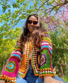 a woman wearing sunglasses and a colorful crochet jacket standing in front of trees