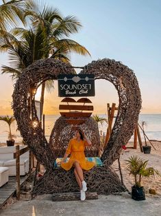 a woman sitting in front of a heart shaped sculpture on the beach with palm trees