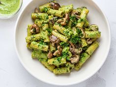 a white bowl filled with pesto pasta and mushrooms next to a green sauce on the side