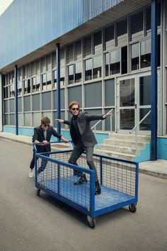 two people riding skateboards on top of a blue cart in front of a building