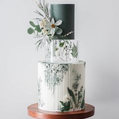 a white and green wedding cake with greenery on the top, sitting on a wooden table