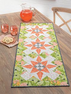 a table with a green and orange quilt on it next to two glasses, a cookie and a teapot