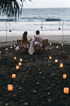 two people sitting at a table on the beach with candles in the shape of hearts