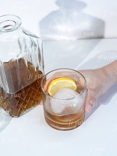 a person holding a glass with ice and lemon in it next to a decanter